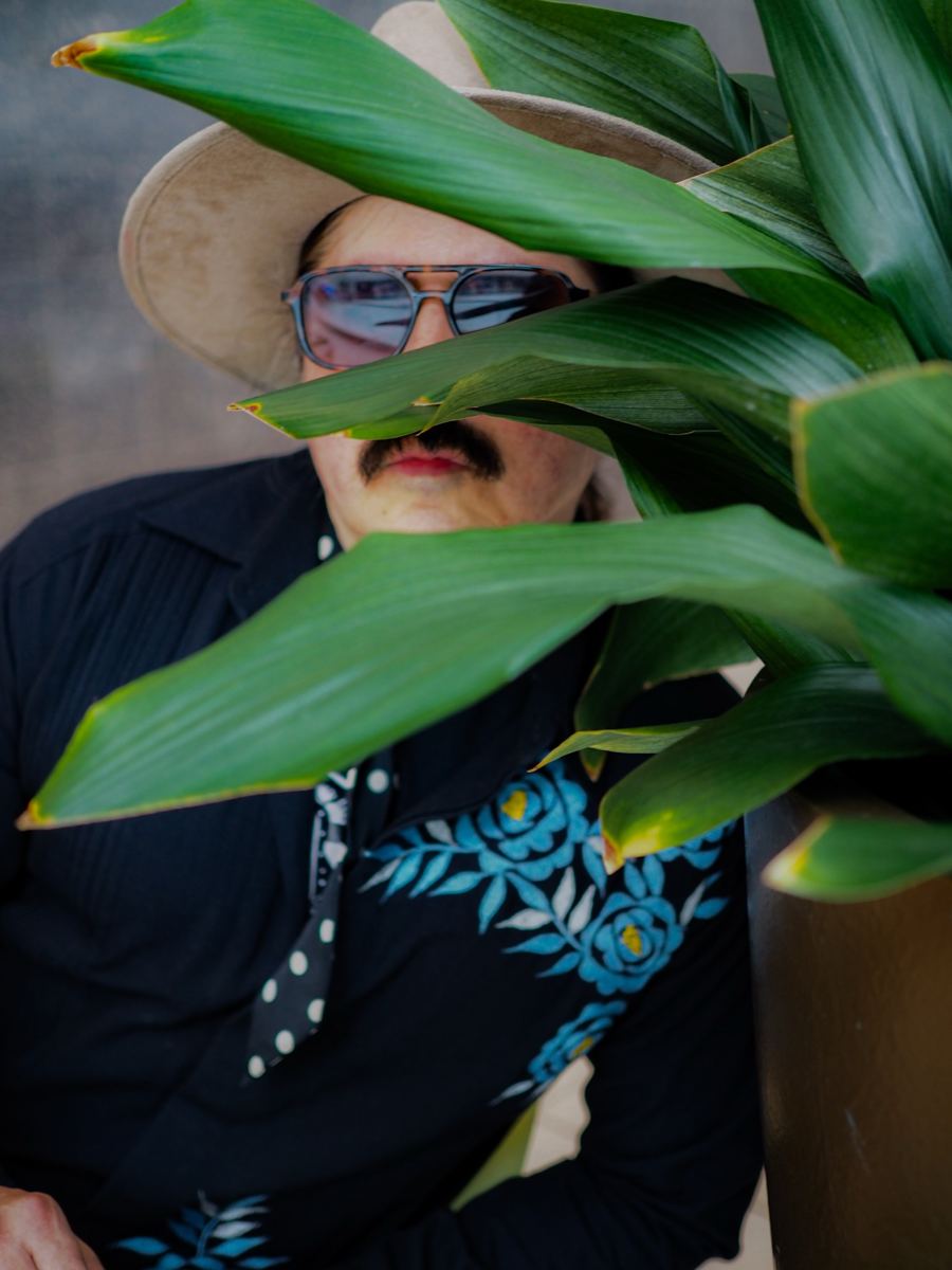 The Ghost of James Jones in a cowboy hat and blue sunglasses, half hidden behind green leaves